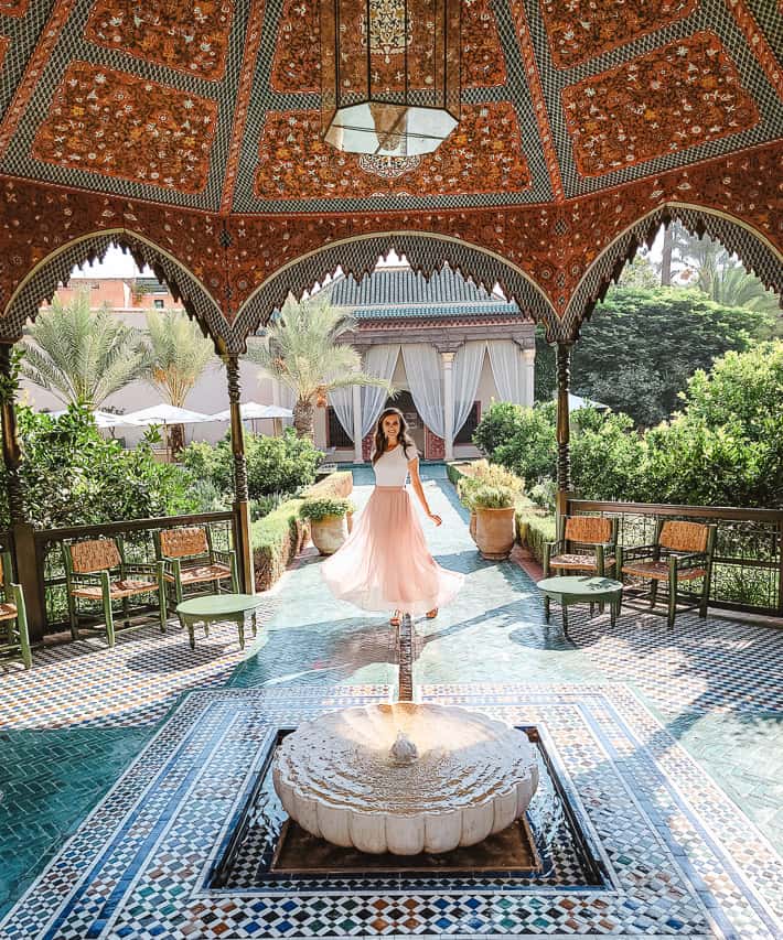 A woman in a long pink skirt stands in the middle of one archway of the gazebo, a low fountain babbling in front of her, tilework on the ground, and arches in the background in the Jardin Secret, one of the prettiest unique things to do in Marrakech.