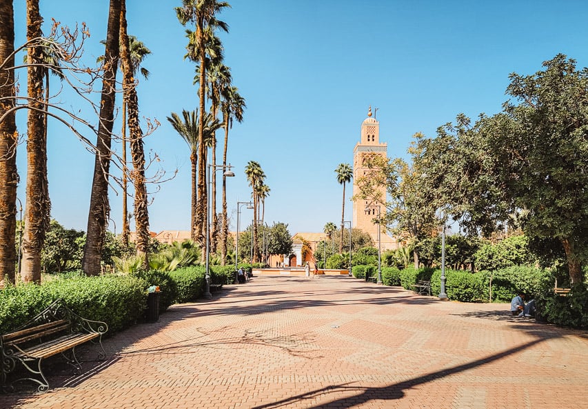 A view of the Koutubia mosque and minaret in Marrakech, with a sidwalk and palm trees in the foreground.