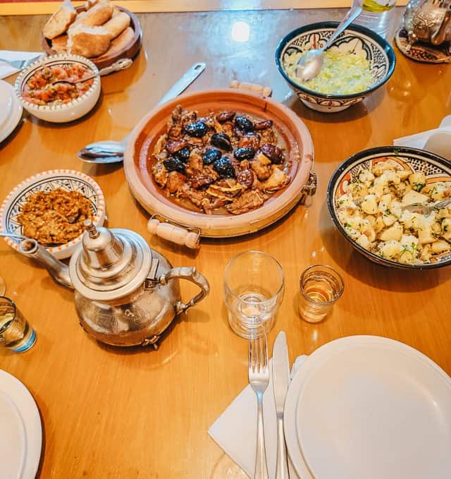 A table is set with white plates, silverware, and cups. A tagine with lamb and apricots is in the center, with a teapot, a bread bowl, and four smaller bowls of salads surrounds it.