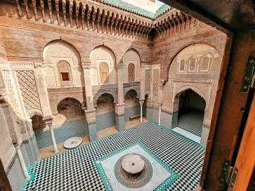 A view of the madrasa (old school) courtyard from a dorm room window on the 2nd floor. You can see arches with carved stucco, columns, a water feature in the middle of the room, green tiles, with the floor being mostly black and white diamond tiles.