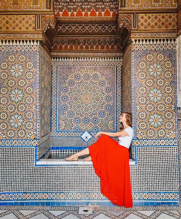 A woman in a white shirt and long red skirt sits in an alcove, looking at the intricate, geometric colorful tiles covering the surface of the entire alcove in the Musee dar el bacha.