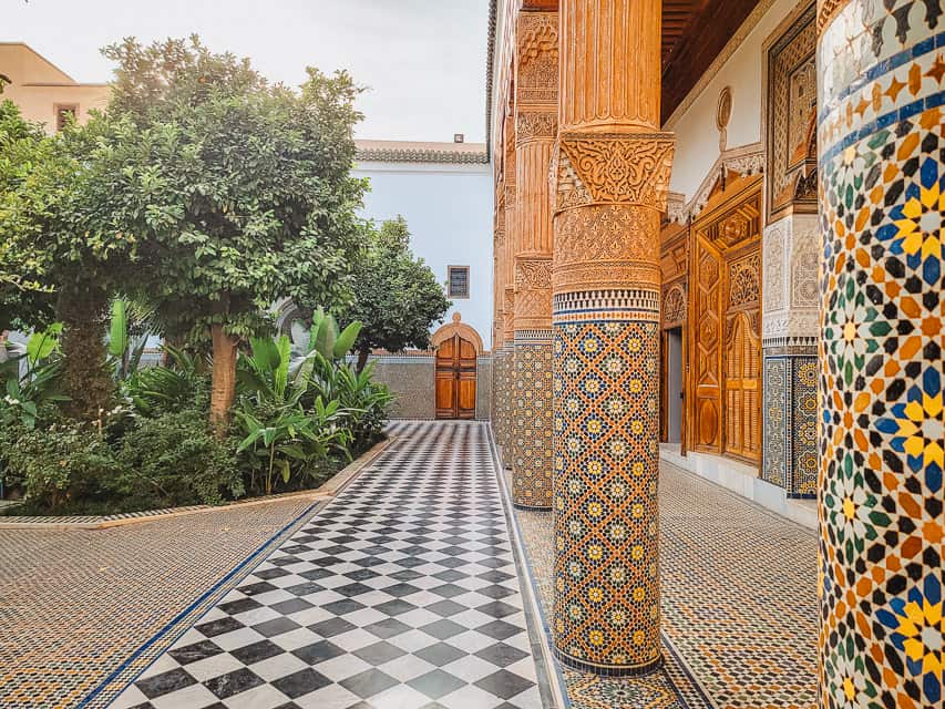 a black and white checkered tile floor extends in front of you, with plants and trees on the left side, and colorful, mosaics on columns on the right side in the Musee dar el Bacha.