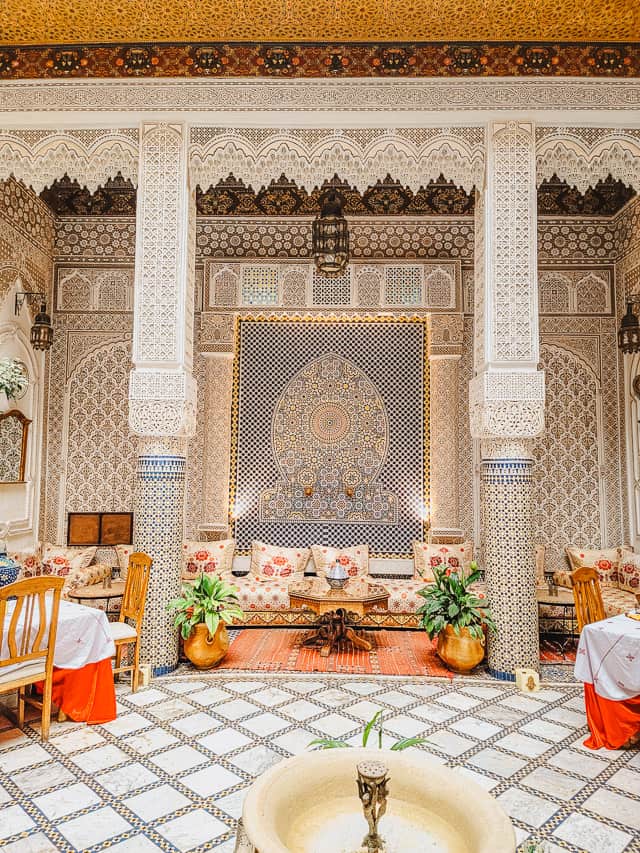 The courtyard of a riad is shown. It is incredibly ornate, with tiles lining the walls and carved stucco and cedarwood ornamentation. There are tables and chairs off to the sides, and a plush bench to sit on lining the walls behind the columns.