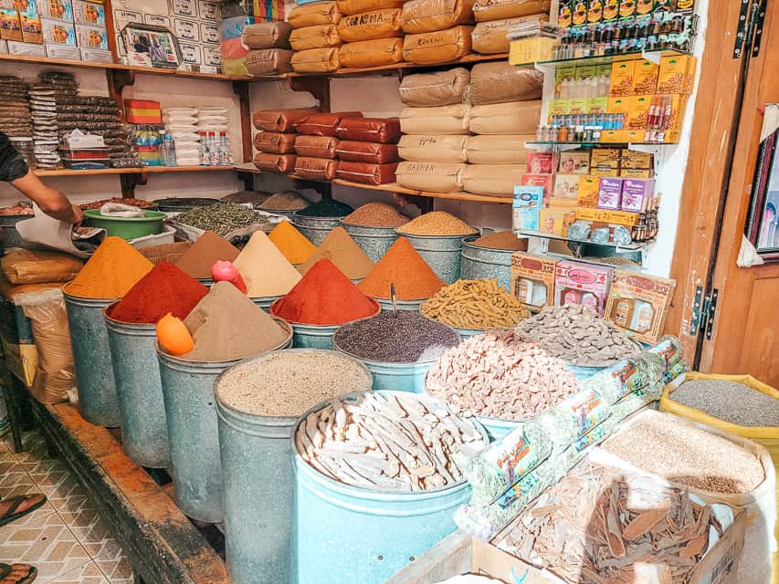 A spice shop is shown. In front are 2 rows of 3 bins each of whole spices (e.g. cinnamon sticks). Behind those are 3 rows of 3 bins each with ground spices, all very colorful (red, orange, white, etc). The spices are mounded into a triangle on top. Behind the bins are shelves stacked with bulk bags of spices.