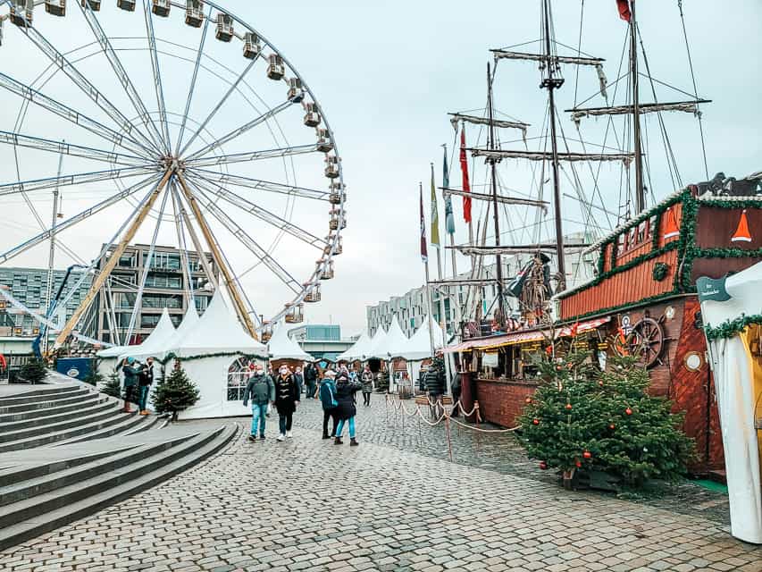 A tall ferris wheel is in the background, and white tents with pointed tops are in the middle ground, and an old timey ship that is selling drinnks is in the foreground.