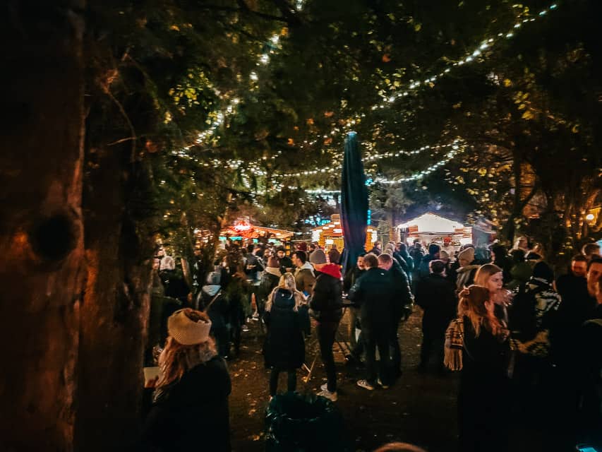 Lots of people gather around bar tables in a park area with lots of trees and a few lights in the Stadtgarden market.