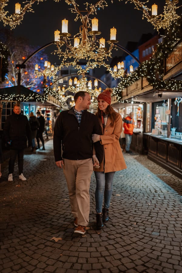 A man and a woman hold hands, smile at each other, and walk down a row of the markets lined with lights and wooden chandeliers.