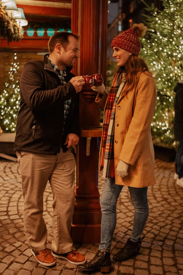 A man and a woman smile at each other, and click their mugs together in the Christmas market.