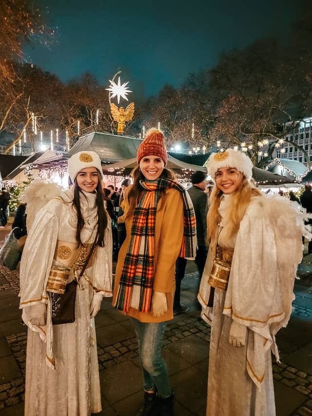 A woman in a beanie, coat, and scarf stands between two women dressed up as angels in the Cologne Christmas market.