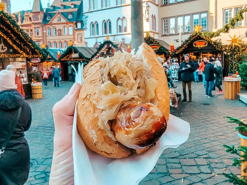A hand holds up a bratwurst in a bun, topped with sauerkraut, with the Trier markets in the background.