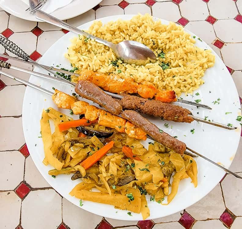 A plate of brochettes (Moroccan kebabs) feature skewers of chicken, beef, and sausage served with veggies and rice.