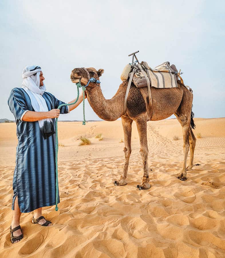 A man in a blue and black kaftan and a white turban scarf looks at a camel in the sahara desert.