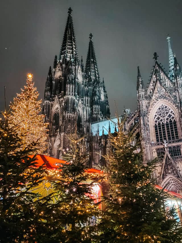 Pine trees covered in twinkling yellow lights are in the foreground, the red roofs of the market stalls in the mid-ground, and the tall, black and white spires of the cologne cathedral in the background, against the night sky.