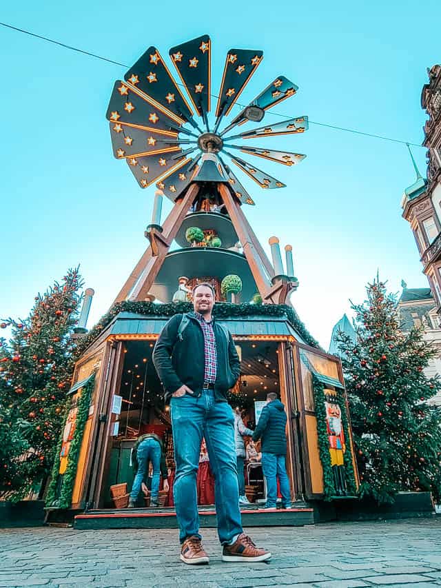 A man stands in front of a Christmas pyramid in Trier. This Christmas pyramid has a small shop underneath, pyramid shaped beams running up to the spinning fan wheel on top.
