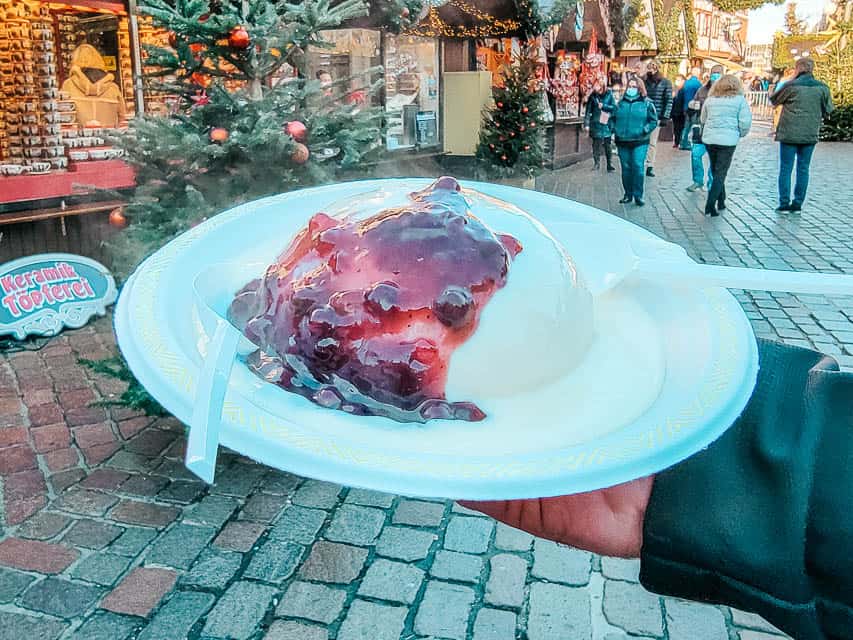 A hand holds a paper plate with dampfnudeln in a market in Trier. This is an oversized rounded dumpling with vanilla sauce and fruit sauce on top.