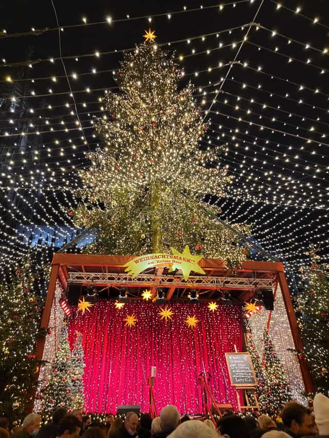 A giant pine tree decorated with thousands of lights stands tall above a stage hung with red curtains. A web of lights extends from the tree above the stage, covering the surrounding area.