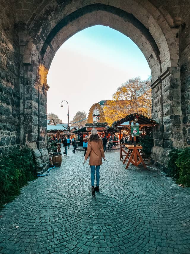 A woman in a coat and white beanie walks through a grand, stone arched entrance to St. Nicolas Market.