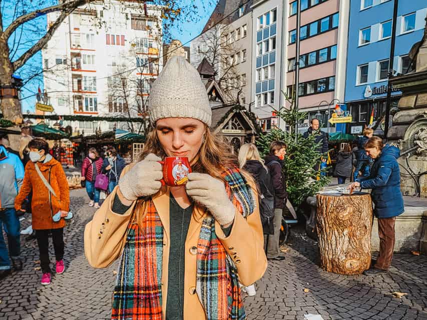 A woman in a coat, white hat, and scarf sips from a red mug in the middle of a market during the day.