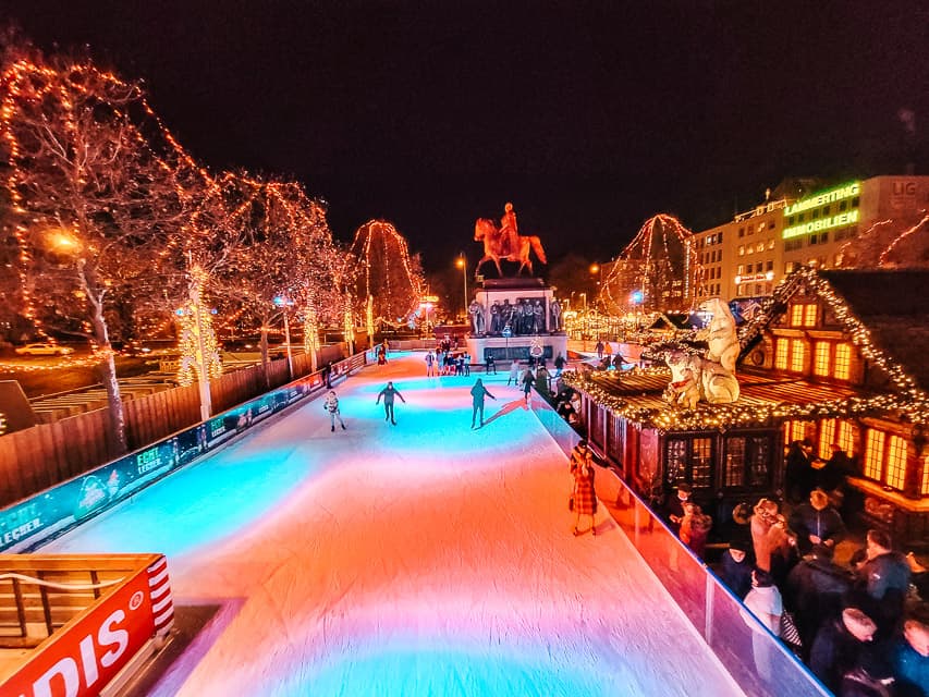 A look from above of the ice skating rink in Cologne - with a few skaters on it. Trees and stalls filled with lights line the rink.
