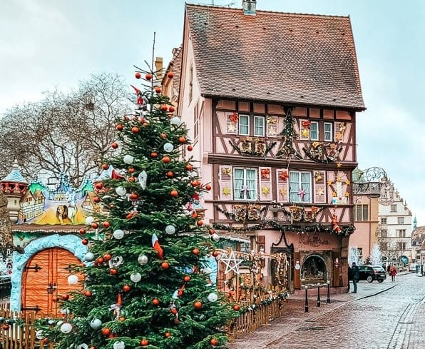 A charming Christmas market scene in Colmar, France. A large Christmas tree stand before a pink, decorated half-timbered home.