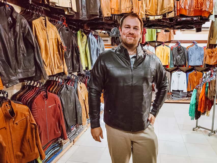 A handsome man looks DANG GOOD in a soft, buttery leather jacket in a leather shop in Fes. Hangers full of leather jackets surround the man. Laethers goods are one of the best things to buy in Fes, Morocco.