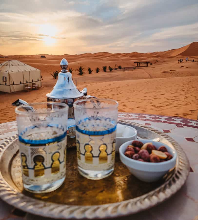 A silver tray with glasses and a teapot rest on a table with orange sand dunes and a sunset in the distance.