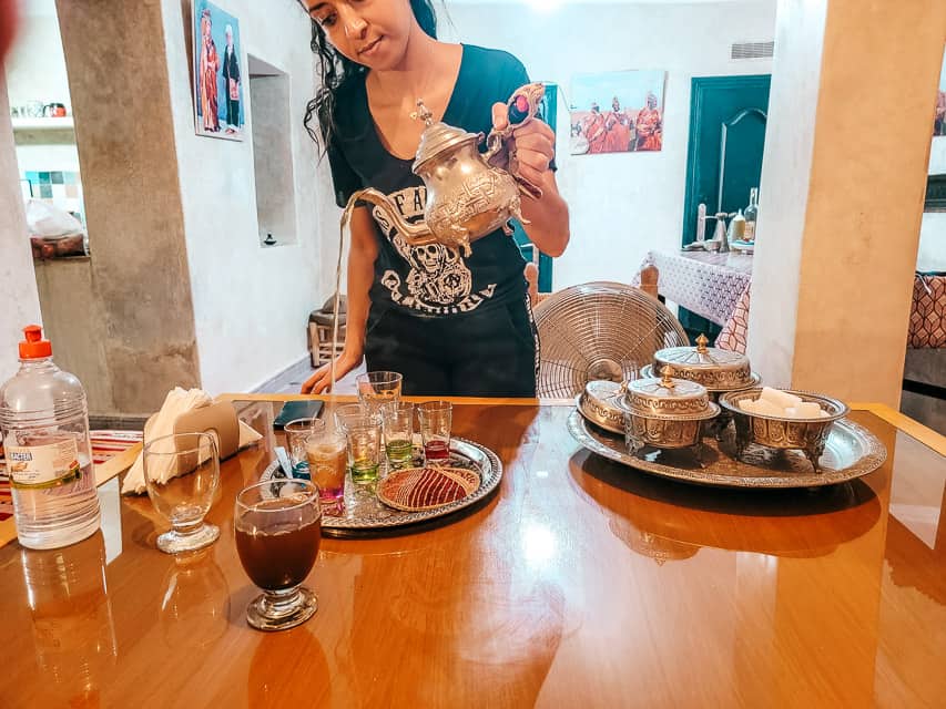 A woman pours mint tea in a very long stream. A tray with covered dishes of things to add, such as sugar is next to her.