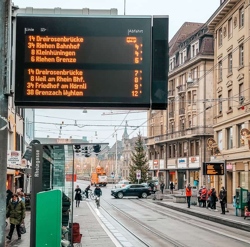 A picture of a regular street in Basel at Christmastime, near a bus stop/tram stop, with a digital sign displaying all the upcoming trams.