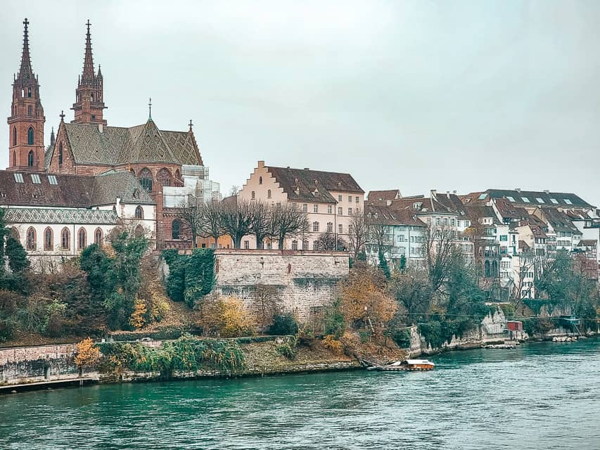 A view of Basel from the Rhine River. You can see the river in front and charming buildings lining the banks of the river, with the spires of the cathedral in the back.