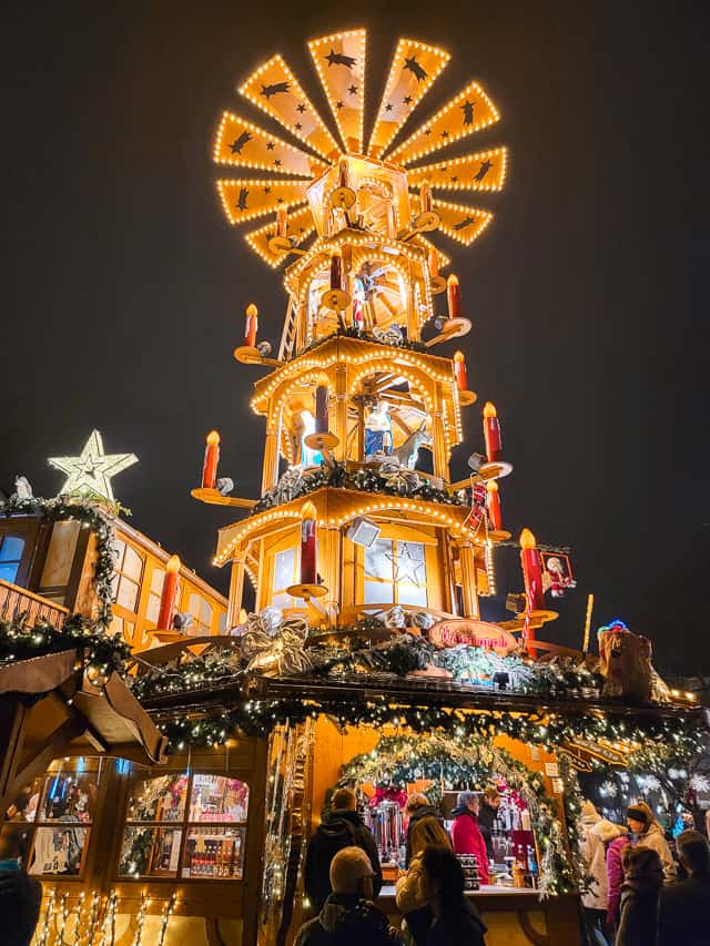 A traditional Christmas pyramid, which is several stories high. 4 different levels are stacked on top of each other, with "fan blades" on the top. Candles adn different figurines are on the tower. The pyramid sits on a top of a little, wodden drink stand, that is trimmed with greenery.