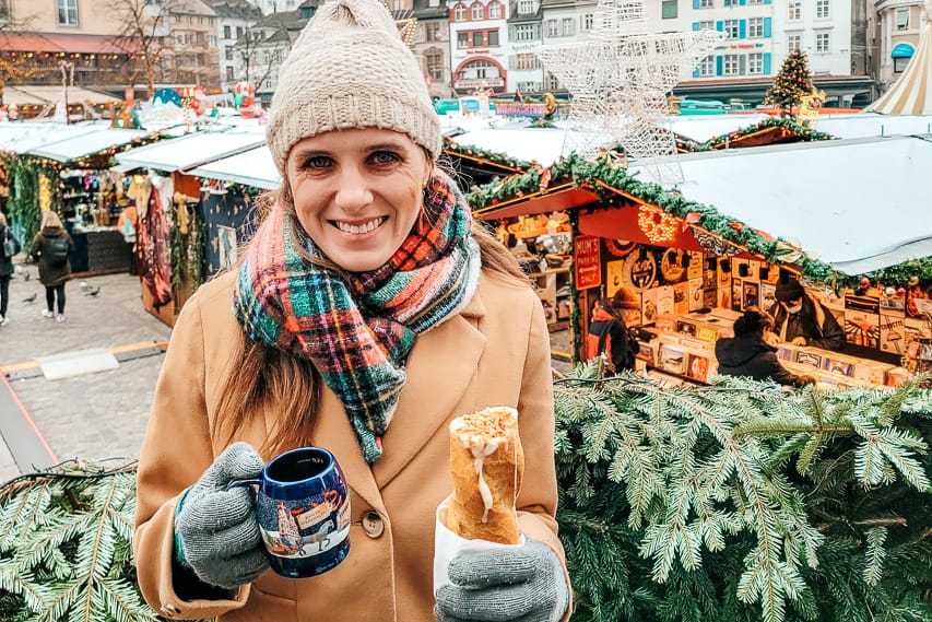 A woman smiles at the camera, holding a blue mug in one hand and a baguette stuffed with cheese in the other - one of the best parts of the Basel Christmas market. Stalls of the Christmas market are set up behind her.