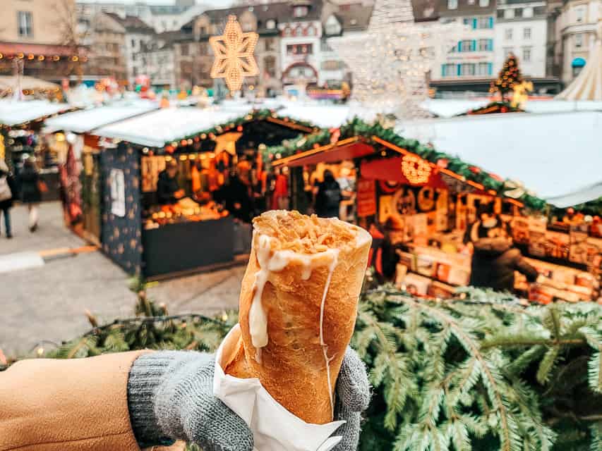 A hand holds a baguette stuffed with fondue in front of the stalls of the Basel Christmas market.