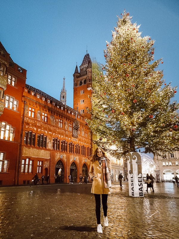 A woman in a tan coat and red scarf smiles at the camera in the middle of a squre with a tall evergreen tree filled with thousands and thousands of lights, and the dark red city hall building behind her.