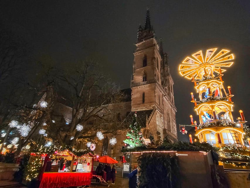 A lit up Christmas pyramid shines against the night sky, with some market stalls in the foreground, lit snowflakes in the trees, and the cathedral in the background.
