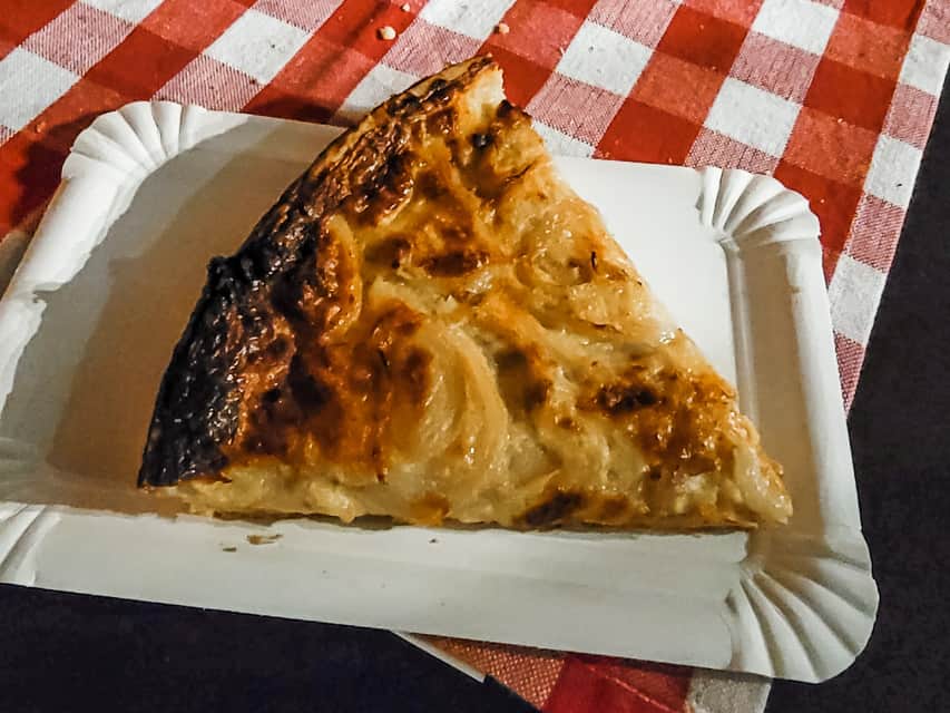A paper white tray on a red and white checked table cloth holds a wedge of quiche lorraine - with extra onions on it.