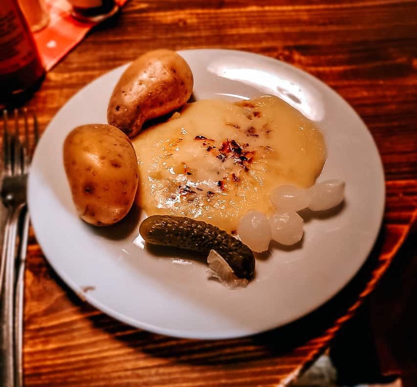 A white plate holds 2 boiled potatoes, a mouind of melted raclette cheese, a pickle, and 4 small pickled onions. This is a plate of traditional raclette.
