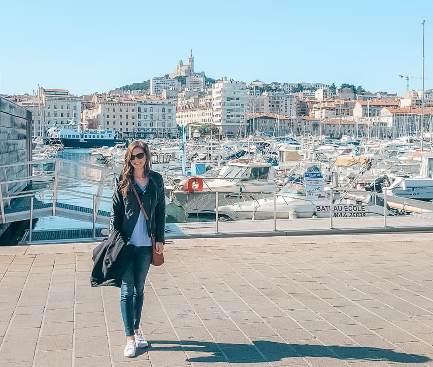 A woman in a leather jacket stands on the sidewalk next to the port with lots of boats and buildings in the background.