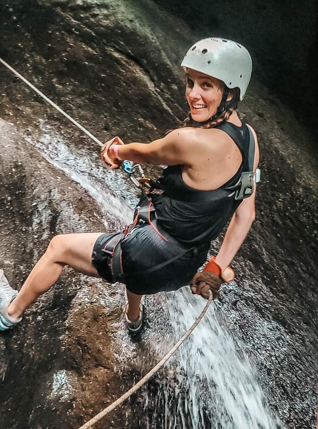 A woman in black is wearing a white helmet and a black harness and is hooked up to a rope, backing up down a rock with water streaming down it.