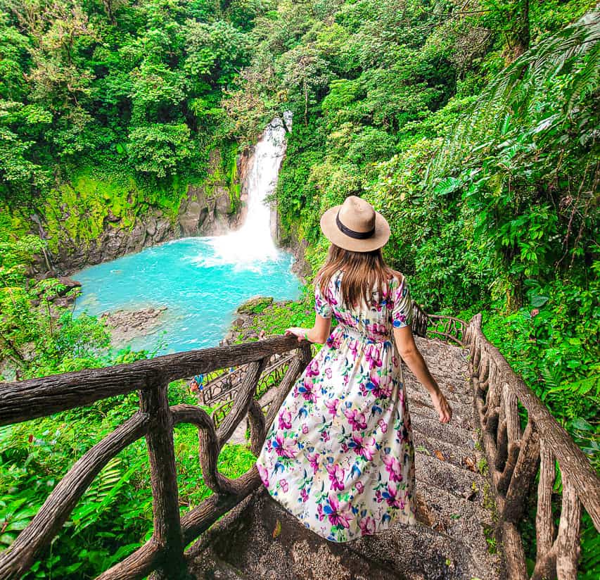 A woman in a flower dress stands on a stairway in the rainforest, looking at a bright blue waterfall, surrounded by thick green foliage.