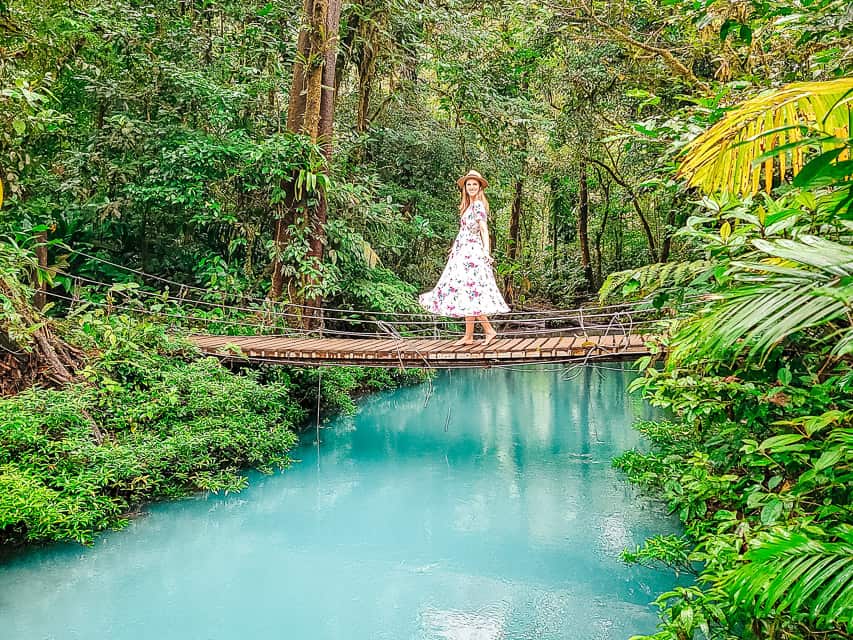 A woman in a white flower dress stands on a wooden bridge over blue water in the rainforest.