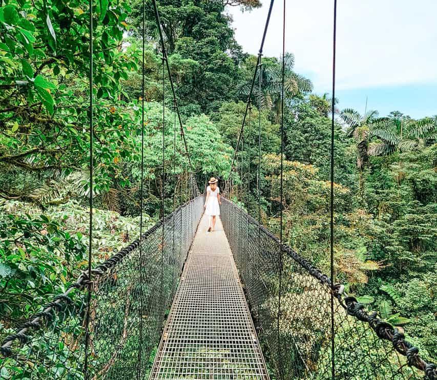 A woman in a white dress and a hat walks down a hanging suspension bridge in the rainforest - one of the best things to do in La Fortuna.