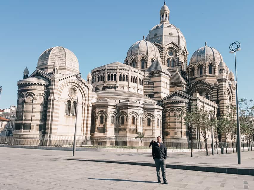 A striking large cathedral with lots of colorful lines and domes on the edge of the sidewalk with trees lining the edge of the cathedral.