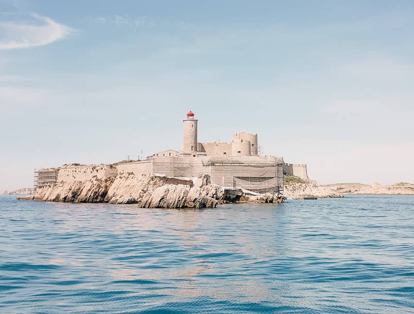 A stone fortress on an island in the middle of Mediterranean Sea with blue water and white limestone land in the background.