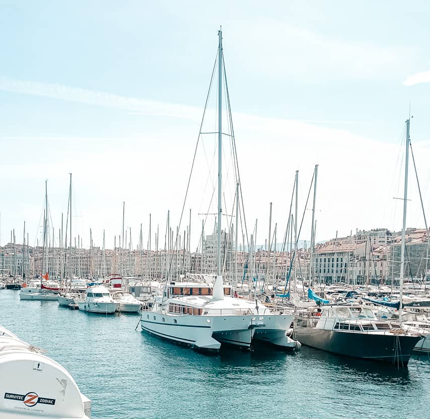 A port in Marseille with deep blue water with hundreds of boats and the outline of stone buildings in the background.