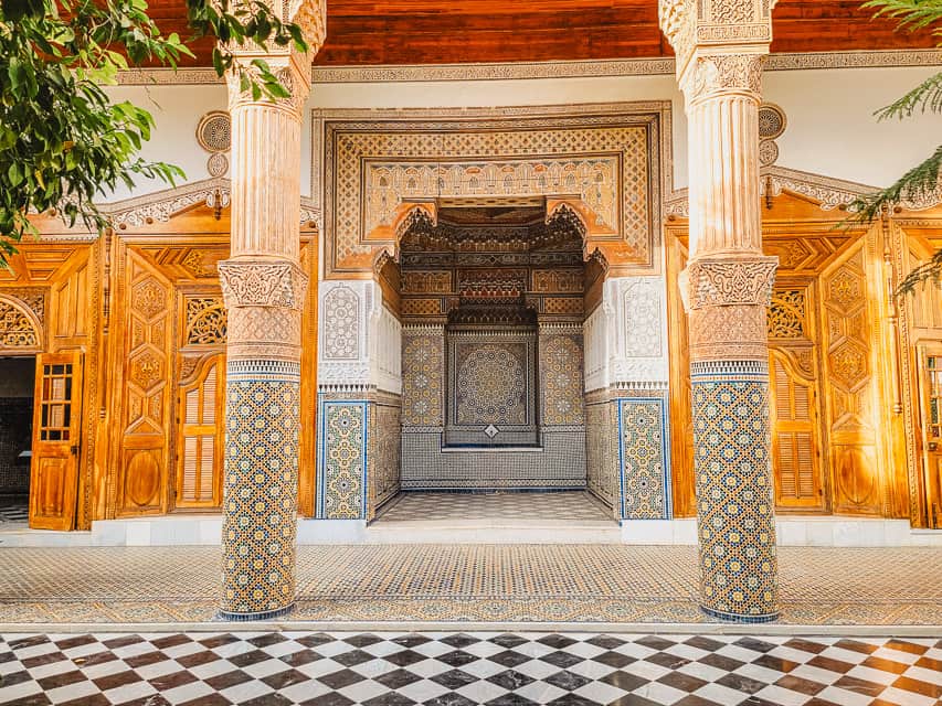 An incredibly beautiful garden room with heavy, detailed tilework, white carved stucco, black and white tile flooring, and dark wood accents in the Musee dar el Bacha.
