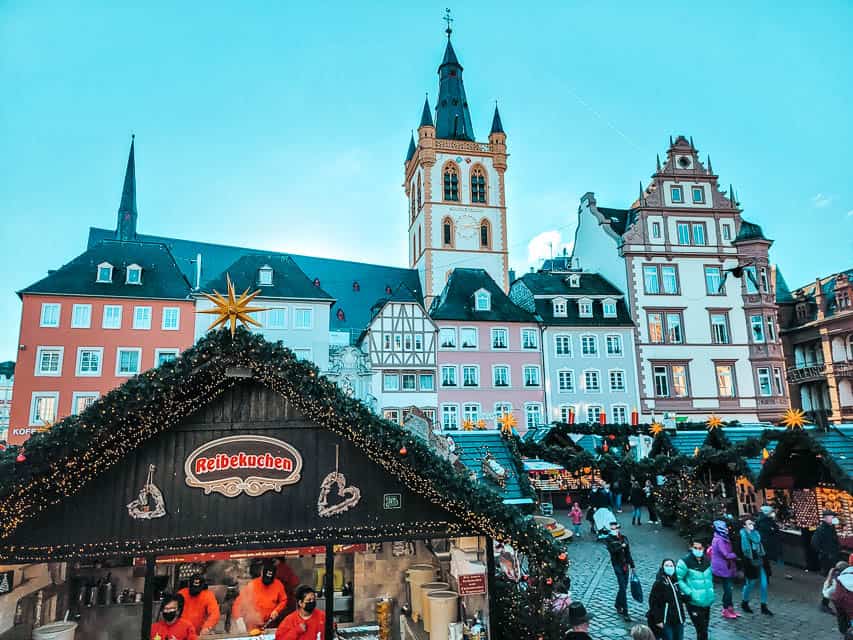 In the foreground: market stalls with pointed roofs and lined with greenery, in the background: the colorful, decorated buildings of Trier. 