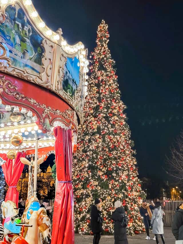 A beautiful carousel is in the front right foreground, with a massive Christmas tree decked out in yellow lights and red bulbs behind it.