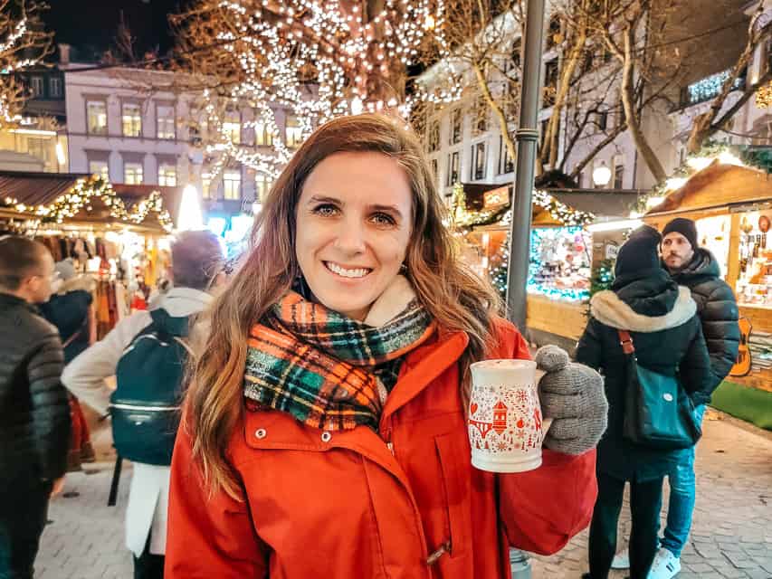 A woman in a red coats smiles at the camera while holding up a white and red mug filled with hoto chocolate. Market stalls and trees filled with lights are behind her.