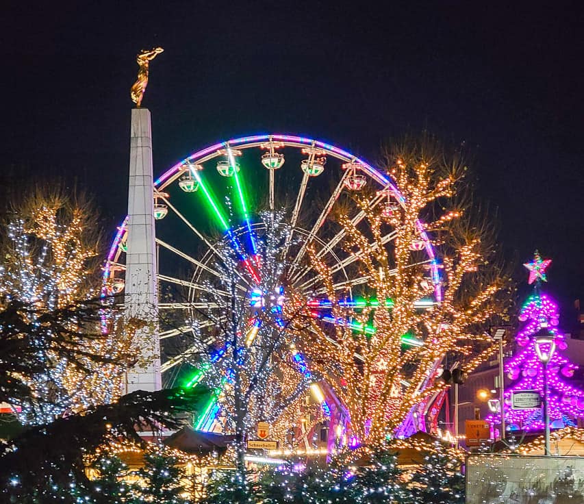 One of the main markets in Luxembourg at the Place de la Constitution, with a tall pillar, grand ferris wheel, games, and trees lit beautifully with thousands of lights.