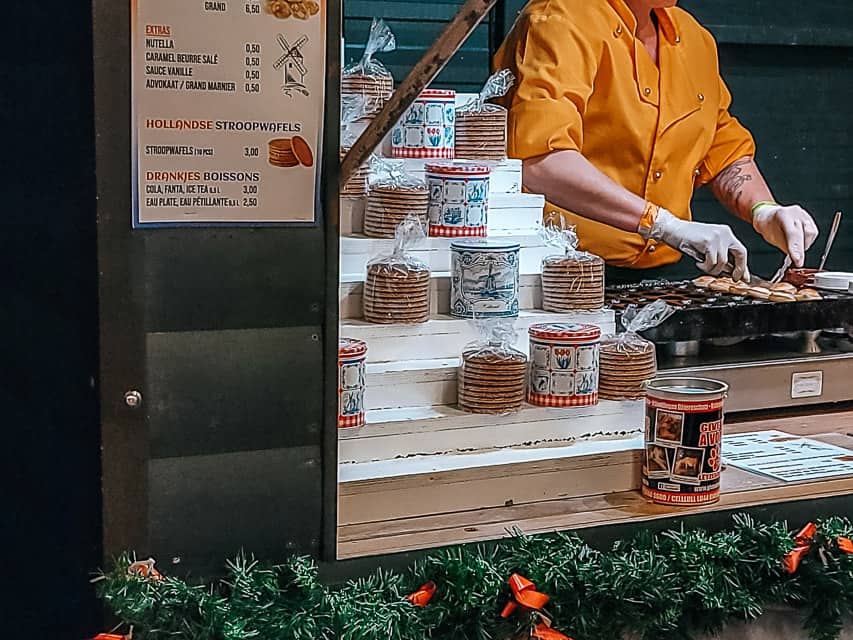 Bags and tins of stroopwaffles are displayed in a stall, and small bite-sized pancakes called pfannkuchen are being made.